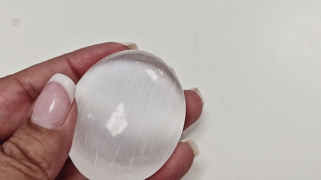 Short video of a 50 mm Selenite Palm Stone being held and rotated in hand, showing its polished oval shape and luminous striations.