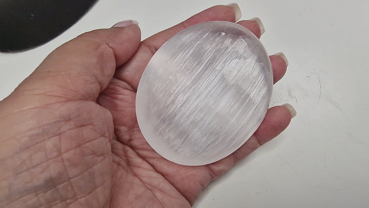 GeoFossils 14-second video of a 70 mm Selenite Palm Stone being held and rotated in hand, showing its polished oval shape, luminous glow, and natural striations from all sides.