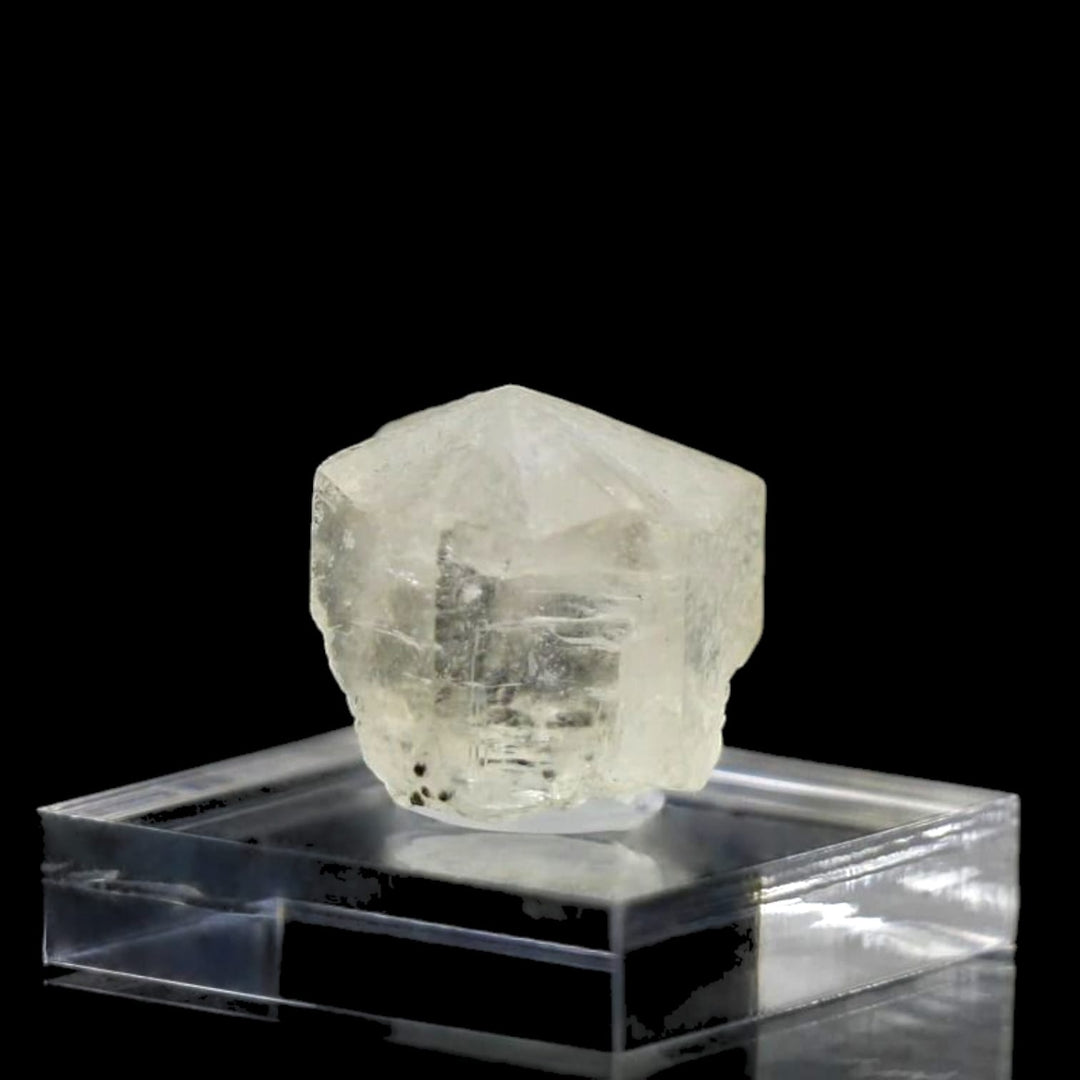 A clear Phenakite crystal displayed on a square glass stand against a black background.