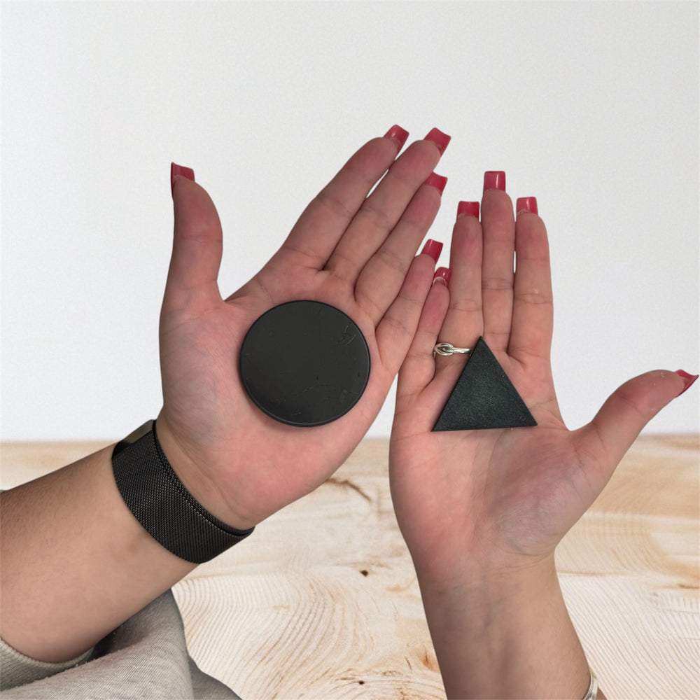 Two hands holding shungite stones for grounding and EMF protection; left hand with round polished shungite crystal, right hand with flat triangular shungite stone.