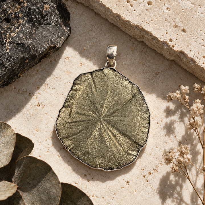 Natural Pyrite Sun pendant styled on warm ivory stone with dried flowers and mineral accents, highlighting its metallic lustre and silver setting.