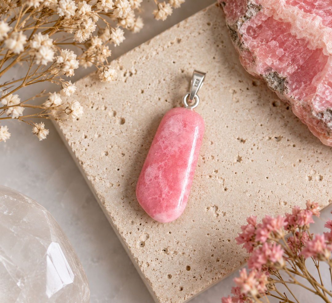 GeoFossils natural Rhodochrosite pendant styled on pale textured stone with delicate dried flowers and complementary pink minerals, highlighting its polished pink gemstone and silver bail.