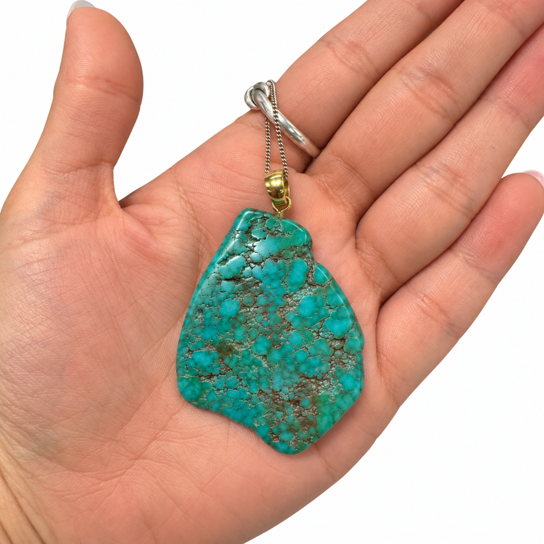 GeoFossils Royston Turquoise freeform pendant resting in the palm of a hand, showing its natural size, asymmetrical shape, intricate matrix and gold-tone bail.