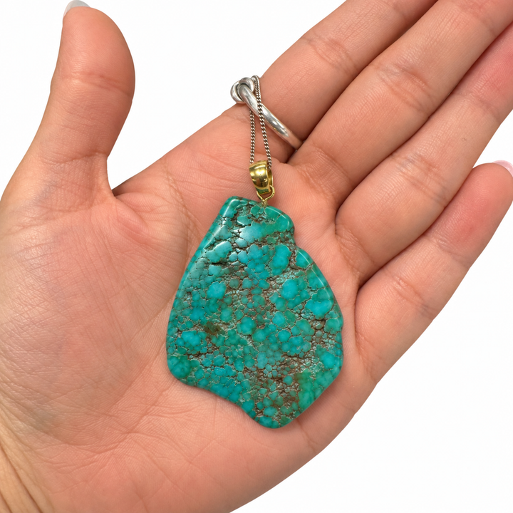 GeoFossils Royston Turquoise freeform pendant resting in the palm of a hand, showing its natural size, asymmetrical shape, intricate matrix and gold-tone bail.