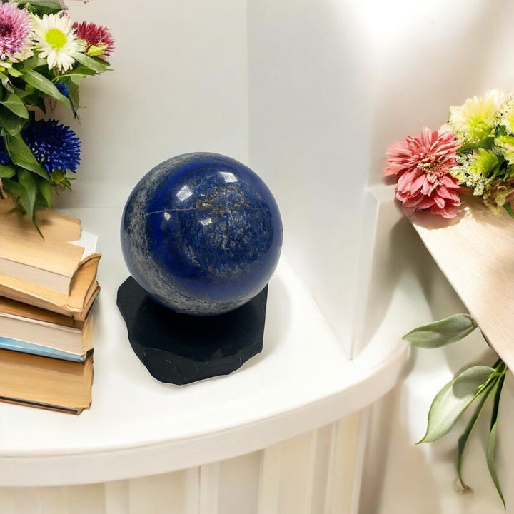Polished lapis lazuli crystal sphere displayed on a natural shungite base, surrounded by books and flowers, used for energy balance, grounding, and EMF protection