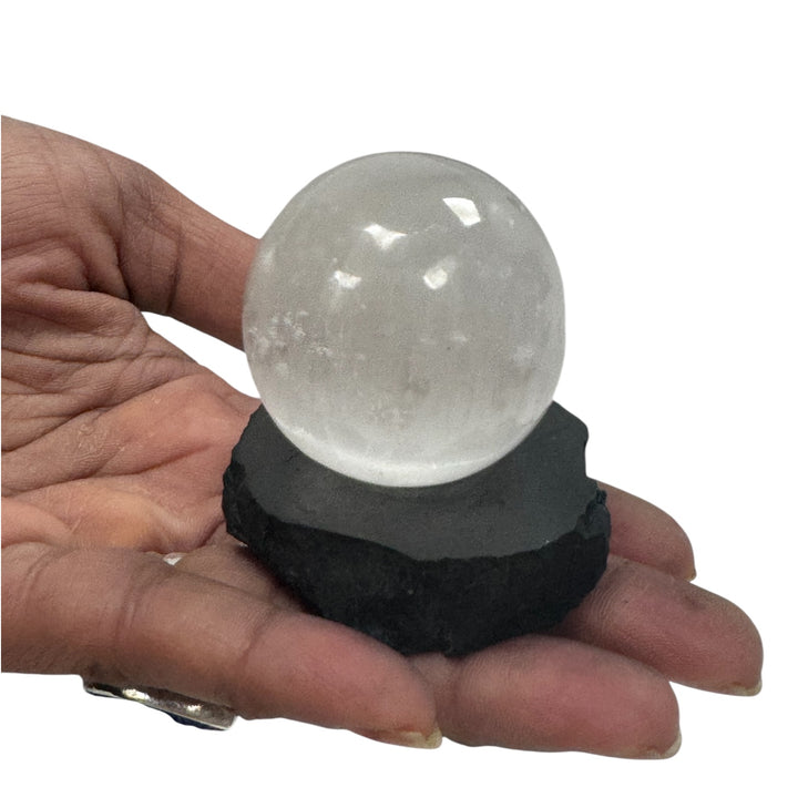 Hand holding a clear quartz crystal sphere placed on a small shungite base for grounding and spiritual cleansing.