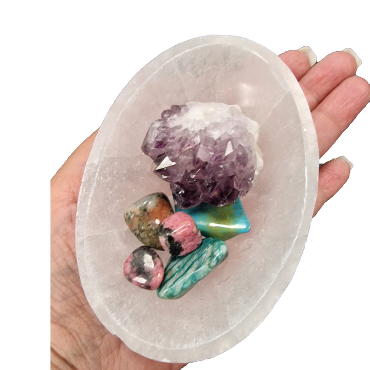 Hand holding a selenite crystal dish with various crystals including amethyst, turquoise, and pink stone.