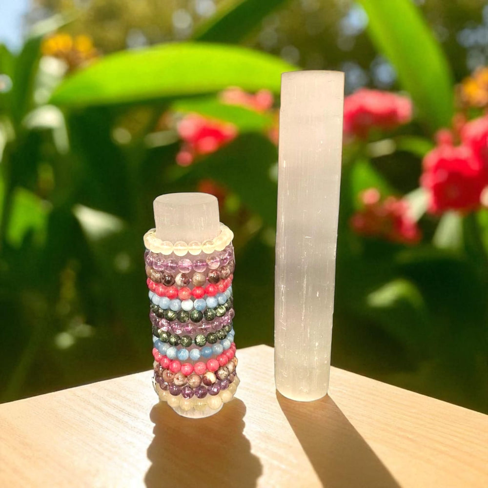 GeoFossils Selenite Bracelet Charging Stand 20cm  and 30cm displayed side by side with a green leaves and red flowers on a pale natural wood table – ideal for energising spiritual jewellery