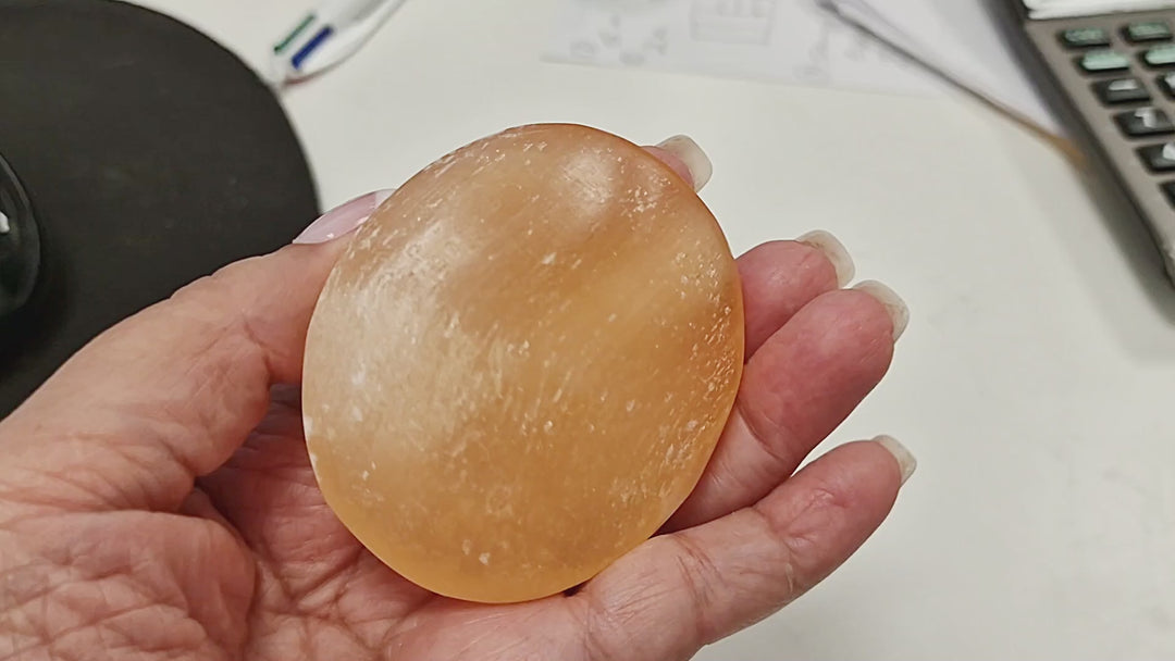 16-second video of a Peach Selenite Palm Stone 70 mm being held and rotated in hand, showing its polished oval shape, warm peach–orange tone, and natural striations from all sides.
