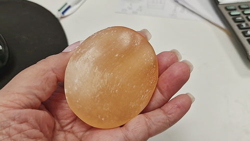 16-second video of a Peach Selenite Palm Stone 70 mm being held and rotated in hand, showing its polished oval shape, warm peach–orange tone, and natural striations from all sides.