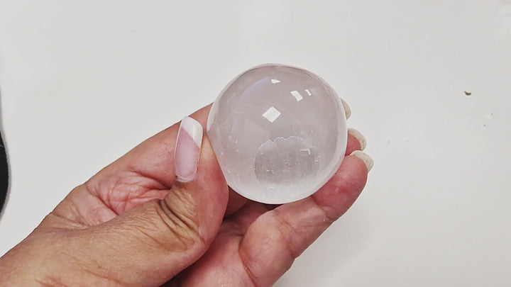 14-second video of a Selenite Sphere 40mm being rotated in hand, showing its smooth polished surface, luminous glow, and natural striations from every angle. Sphere stand used for demonstration but not included.