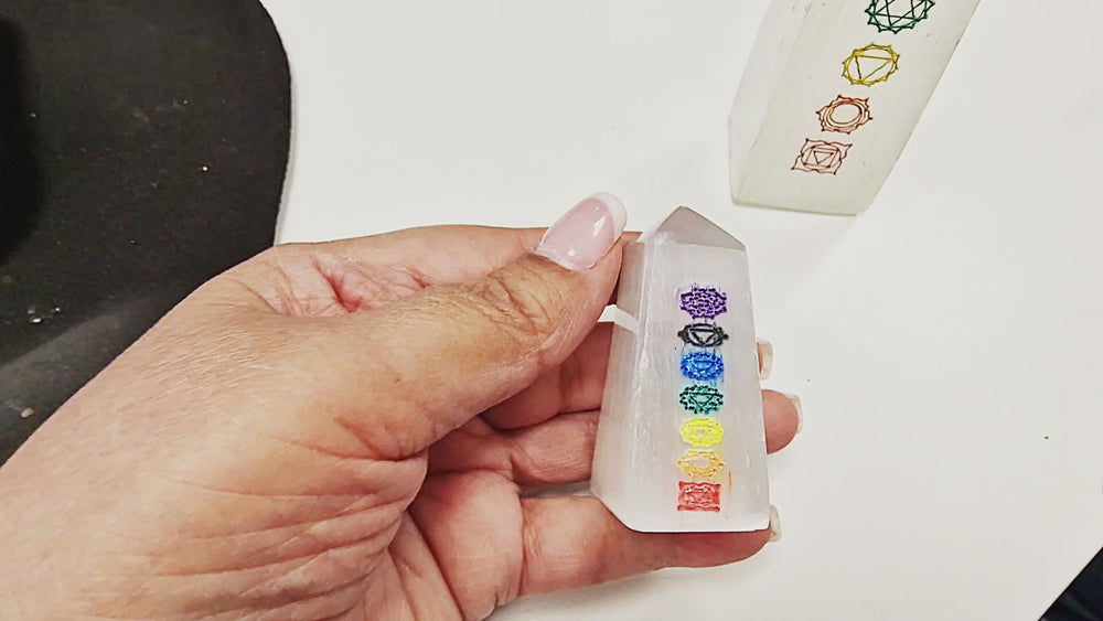 15 second video showcasing two Selenite Chakra Towers, 5cm and 12cm sizes, both polished with 7 chakra symbols engraved. Each tower is shown in hand and rotated to display the colourful chakra engravings and polished surfaces from all angles.